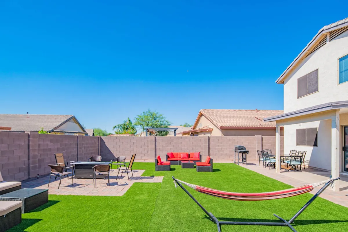 A wide exterior view featuring a red seating area, a family-sized dining table set on a lush green lawn, and a hammock beneath a sunny sky. The atmosphere is casual and inviting, ideal for socializing, outdoor meals, or relaxing with loved ones.