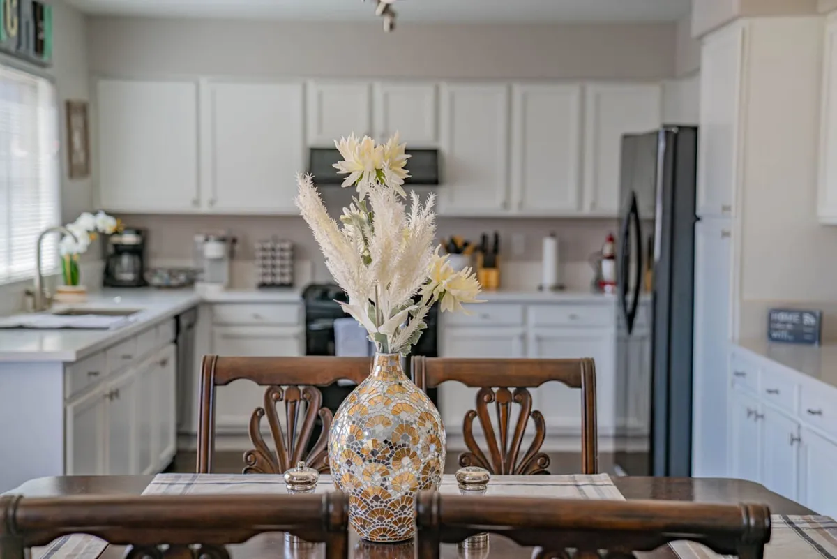 A kitchen table set with a vase of flowers, surrounded by white cabinetry and black appliances in the background. The table suggests a casual place for morning coffee, quick bites, or family gatherings, and the kitchen features all the essentials.