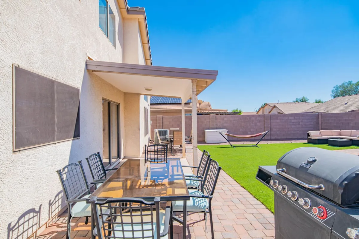 This exterior image highlights a vibrant backyard patio setup, including a large dining table with chairs, a grill for barbecuing, a hammock perfect for unwinding, and a spacious lawn area for games or relaxing in the sun. The overall mood is casual and relaxed, ideal for family gatherings.