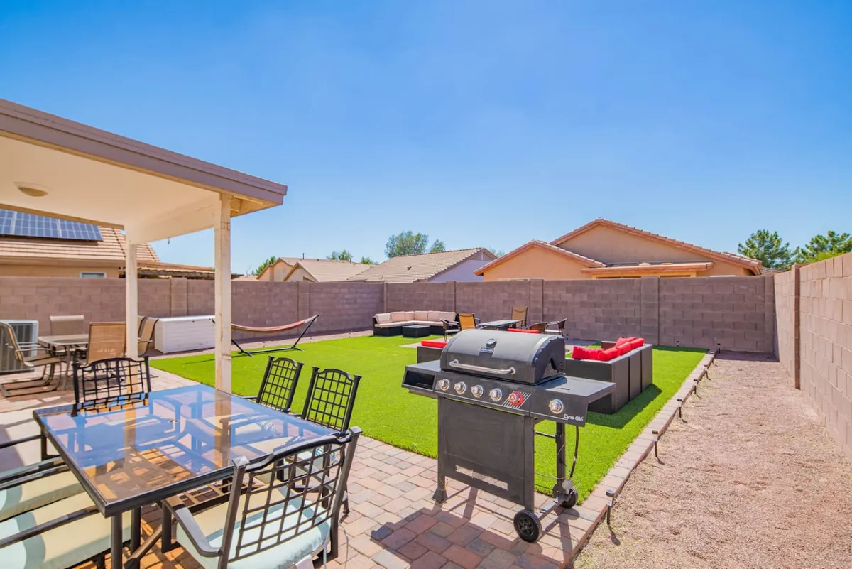 A backyard scene featuring a grill, outdoor seating area, hammock, and dining table. The space is set up for social gatherings, with plenty of room for group activities, barbecuing, and relaxing under the open sky. The ambiance is casual and relaxed, suggesting a perfect spot for evening cookouts or stargazing.