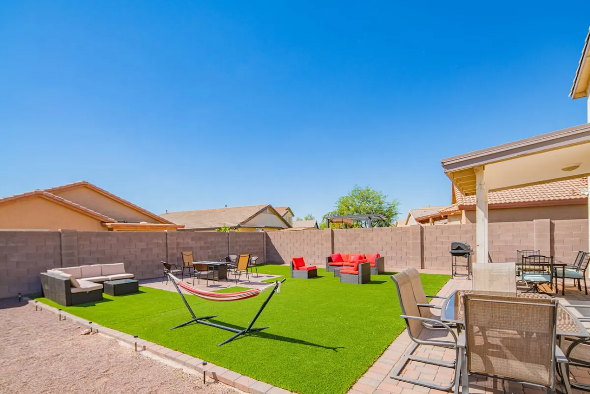 This exterior scene displays a green artificial lawn bordered by patio seating, red accent chairs, an outdoor dining set, and a gently swaying hammock. The space is lively and well-suited for celebrations, games, or quiet relaxation during warm evenings.