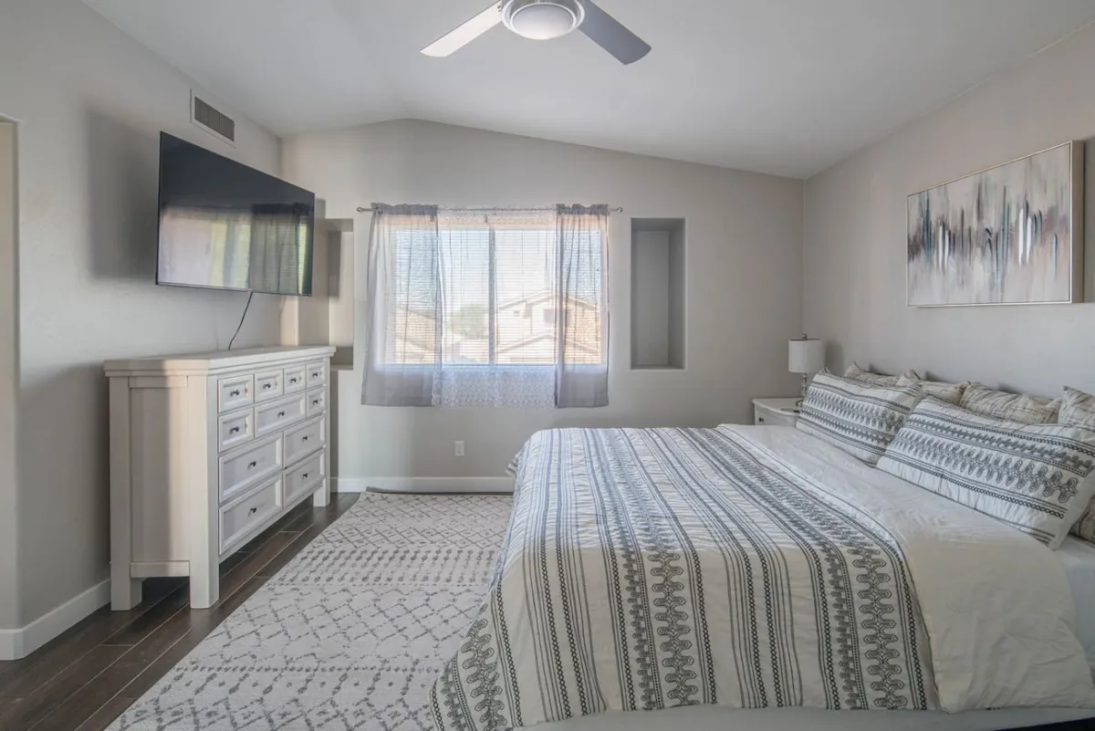 A tranquil bedroom with a bed dressed in patterned bedding, a large wall-mounted Smart TV, a window with sheer curtains letting in soft daylight, and a dresser with decorative accents. The space feels inviting and is tailored for relaxation and personal downtime.