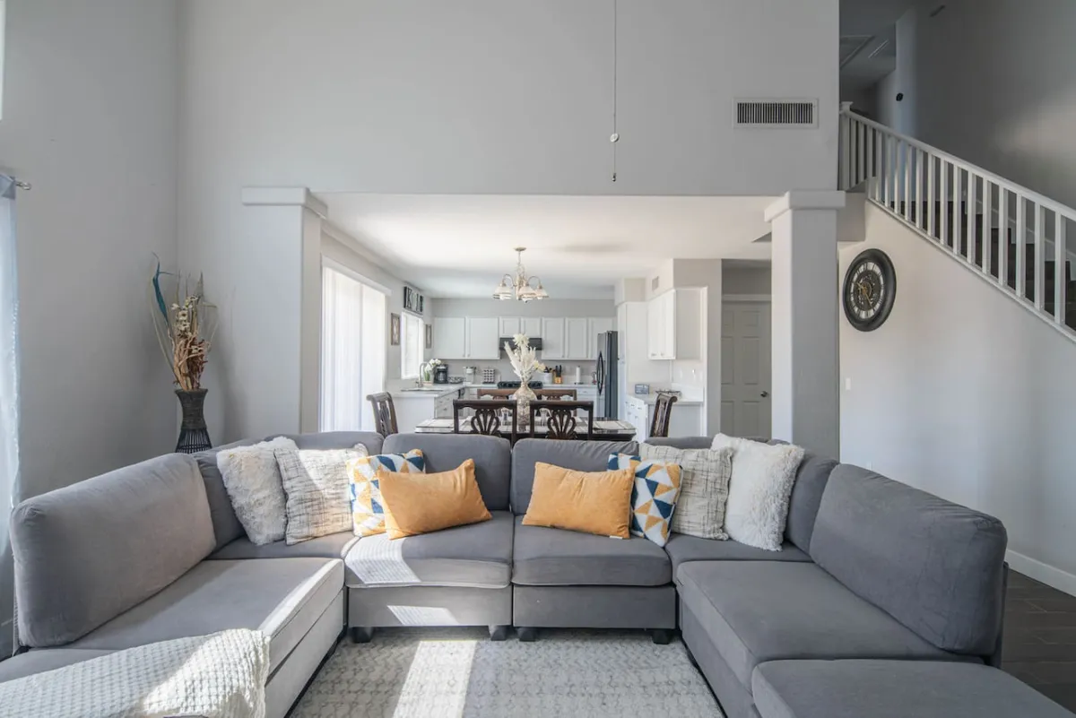 A wide view of the main living area showing a large sectional sofa, a dining area with a table set for six, and the adjacent kitchen. Decorative wall art adds a homey touch. The layout makes it easy for guests to move between relaxing, dining, and meal prep—ideal for both fun and business gatherings.