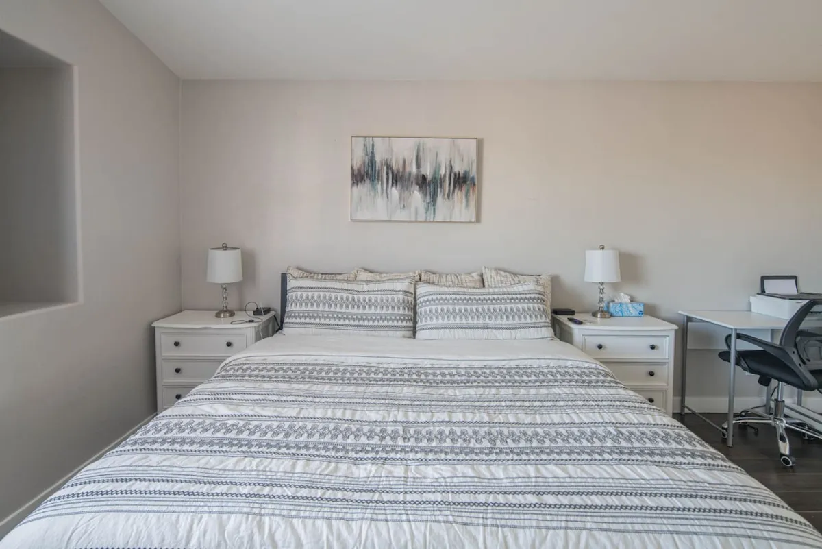 This serene bedroom features a large bed with plush bedding, decorative wall art, and nightstands with lamps. A work desk sits in the corner, offering a functional space for reading or working. Natural light filters in, creating a restful and calm atmosphere ideal for a good night's sleep.