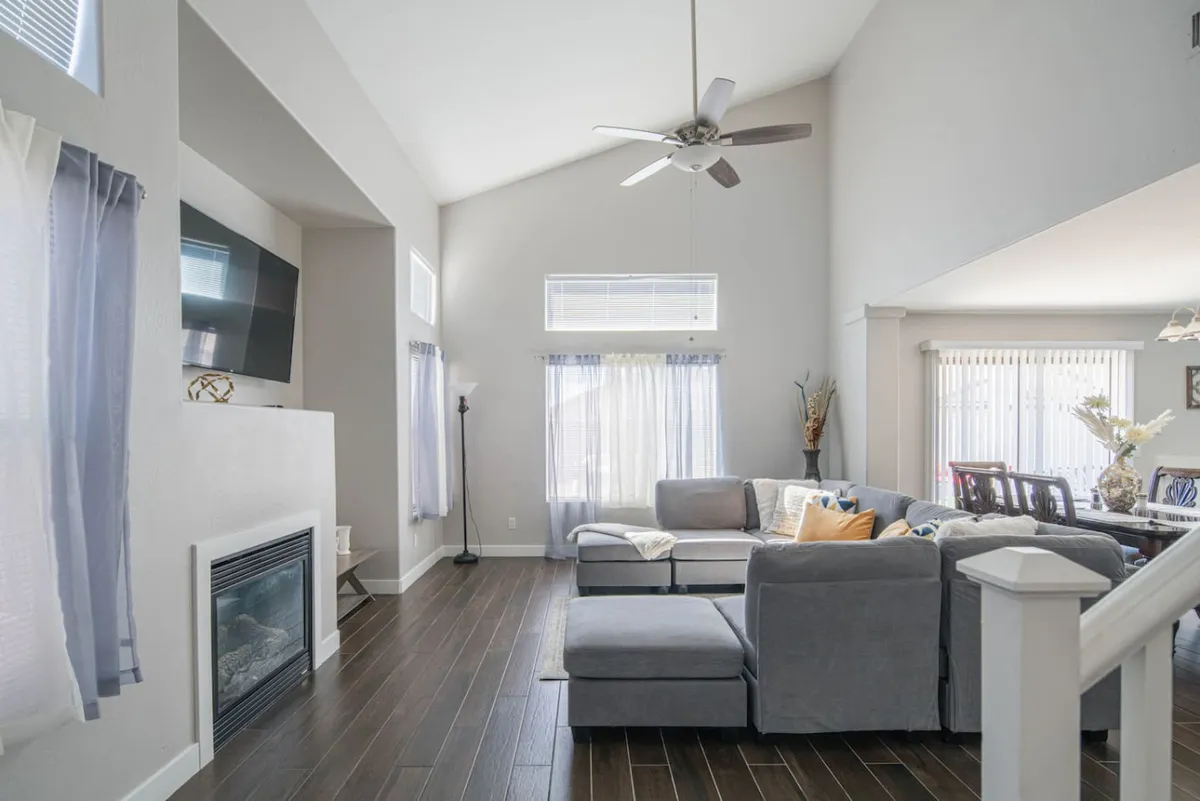 An inviting open-concept living area featuring a large, plush sectional sofa positioned around a fireplace, with ample natural light flooding in through large windows. The dining table sits nearby, offering a seamless flow between lounging and dining spaces. The room is styled for comfort, family gatherings, and group entertainment, with a smart TV visible for movie nights.