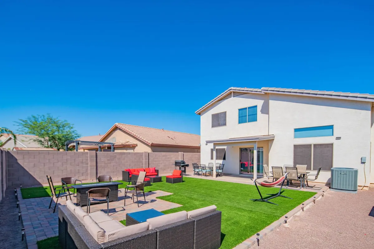 A spacious backyard featuring a green artificial lawn, a cozy red seating area surrounding a fire pit, comfortable patio furniture, and a hammock swung between two supports. The area is designed for entertaining, with plenty of room for lounging or socializing under open skies.