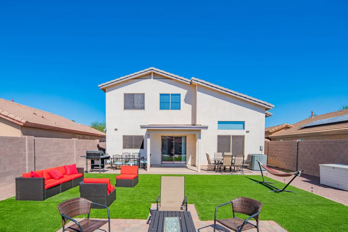 The image shows a vibrant backyard with a lush artificial green lawn, a cozy red outdoor seating area, a hammock for lounging, a grill ready for barbecues, and inviting patio furniture—all set for gatherings and relaxation under the open sky.